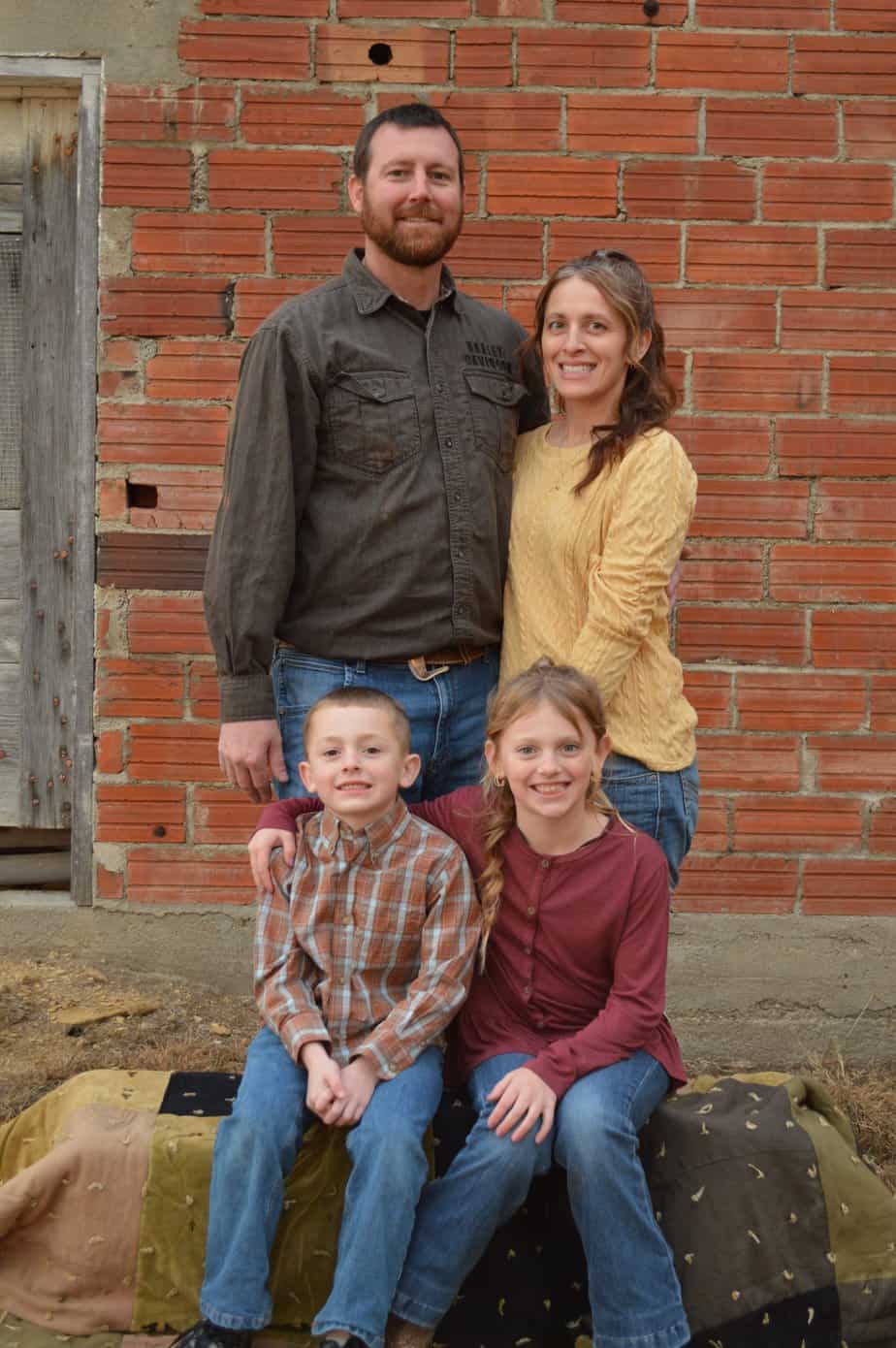 A family of four posing happily in front of a brick wall. the parents stand behind two children, who are sitting on a hay bale, all smiling at the camera.